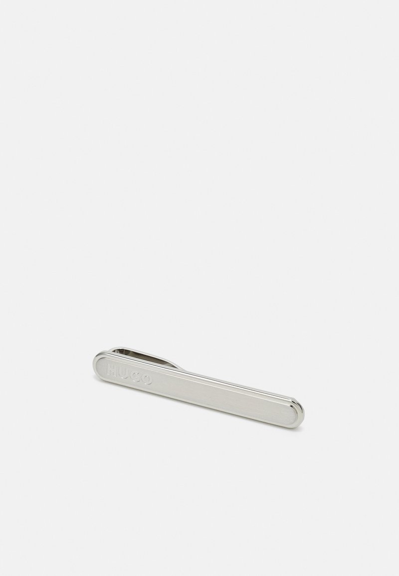 Pince à cravate en métal argenté, au design slim et rectangulaire avec des bords arrondis. Nom de la marque "HUGO" gravé sur un côté. Texture lisse et polie.