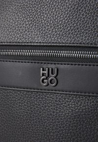Zwarte gestructureerde leren tas met een zilveren rits en een zwart leren accent met het merklogo "HUGO" erop geperst.