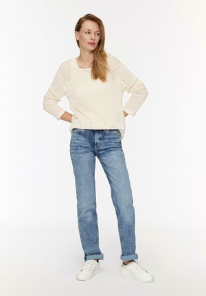 Cremfarbener Strickpullover mit strukturierter Musterung, kombiniert mit hellblauen, geraden Jeans. Träger mit weißen Sneakers.