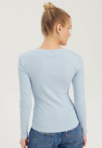 Kvinne med blondt hår i en knute, iført en lys blå ribbet langermet topp og blå denimjeans, sett bakfra.