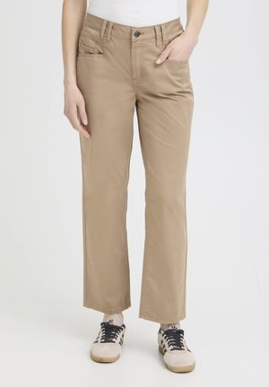 Person iført beige bukser med lige ben, hvid top og hvide sneakers med sorte striber, stående med den ene hånd i lommen.