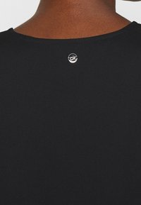 Camisa negra con una textura suave. Presenta un escote redondo y un pequeño logo plateado en la parte posterior cerca del cuello.