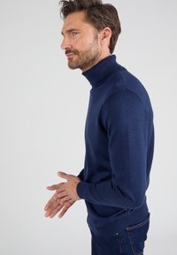 Suéter de cuello alto en azul marino, de material de punto suave, diseño ajustado, con cuello y puños de canalé, combinado con jeans azul oscuro.