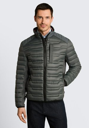 Mann trägt eine dunkelgraue gesteppte Jacke mit Reißverschlusstaschen, steht mit einer Hand in der Tasche vor einem schlichten hellen Hintergrund.