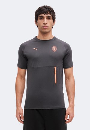 Homme portant un t-shirt Puma gris foncé avec le logo de l'AC Milan et une poche poitrine zippée orange, debout devant un fond uni.