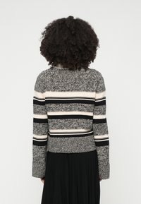 Pleteni pulover z teksturiranim črnim in sivim vzorcem, ki ima široke vodoravne črte v kremni in črni barvi, rebraste manšete in oprijet pas.