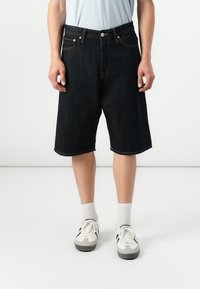 Pantalones cortos de mezclilla azul oscuro con corte recto, diseño de cinco bolsillos, costuras amarillas y dobladillo enrollado, combinados con calcetines blancos y zapatillas.