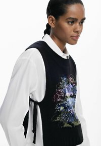 Schwarzer, gestrickter ärmelloser Pullover mit buntem Pailletten-Design, strukturiertem Stoff und verstellbaren Bändern an den Seiten. Getragen über einem weißen Hemd mit Knöpfen.