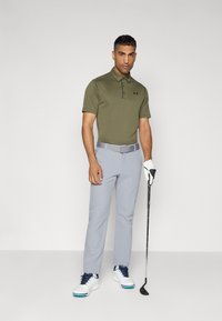 Olivovo zelená polo košeľa s gombíkmi, svetlošedé nohavice, biele golfové topánky s modrými akcentmi, drží golfovú palicu, hladká textúra látky.