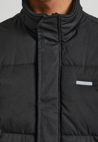 Musta toppatakki, jossa on korkea kaulus, pystysuurella ompelulla ja pieni "Pepe Jeans" -logo edessä. Tasainen kangaspinta.