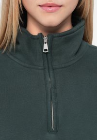 Σκούρο πράσινο φλίς πουλόβερ με ψηλό γιακά και μπροστινό φερμουάρ μισού. Διαθέτει επίπεδες ραφές και λεία, απαλή υφή.