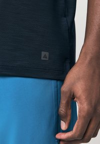 Schwarzes sportliches Shirt mit strukturiertem Stoff, versehen mit einem kleinen grauen Logotag am Saum. Hand hält nahe einer blauen Sportshorts.