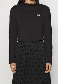 Svart kortärmad topp med rynkad halslinning och broderad vit logotyp. Matchas med en svart kjol med stjärnmönster.