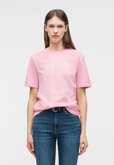 Νεαρή γυναίκα που φοράει ανοιχτό ροζ μπλουζάκι με το μήνυμα "Paris Amour" και μπλε τζιν, στέκεται με τα χέρια στα πλάγια μπροστά σε απλό φόντο.