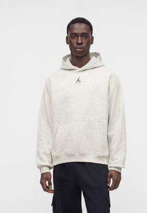 Mężczyzna ubrany w jasnoszarą bluzę z kapturem z małym czerwonym logo Air Jordan na piersi oraz czarne spodnie bojówki, stoi na jednolitym tle.