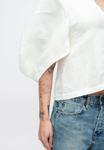 Λευκό cropped τοπ με φουσκωτά μανίκια, φτιαγμένο από ελαφρύ ύφασμα· συνδυάζεται με σκισμένο μπλε τζιν παντελόνι. Ορατά τατουάζ στο χέρι.