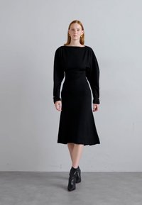 Black long-sleeve dress with a fitted bodice and flared skirt. Smooth fabric; features silver accents at the sleeve cuffs. Worn with black ankle boots.