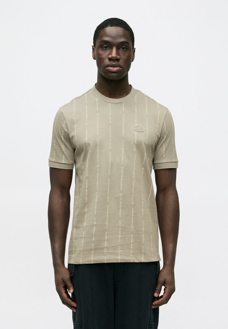 Camiseta beige de manga corta con cuello redondo, con un patrón repetido de logos en un tono ligeramente más oscuro, confeccionada con tela suave.