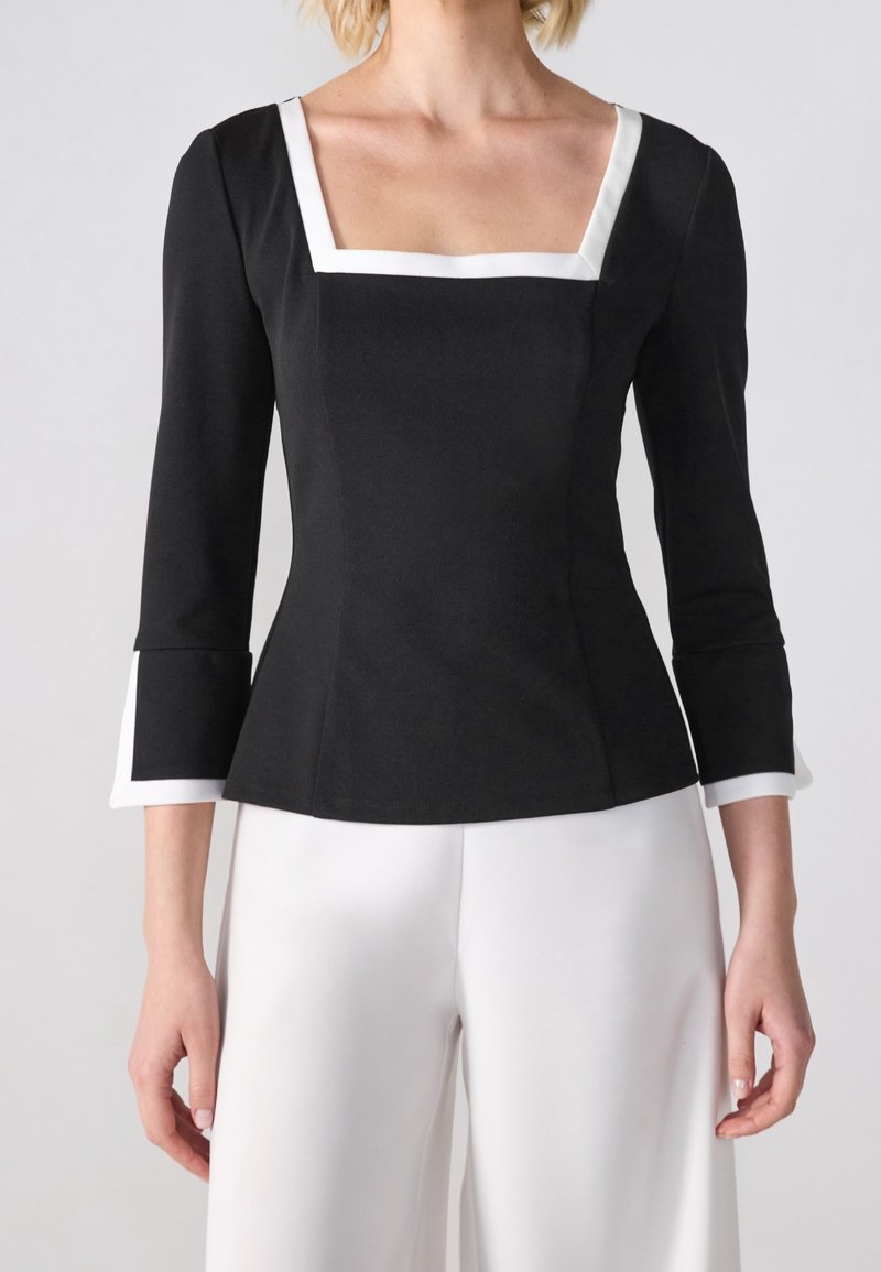 Haut noir ajusté avec un décolleté carré, des manches trois-quarts, et un bord blanc sur les manches et le col. Texture de tissu lisse.