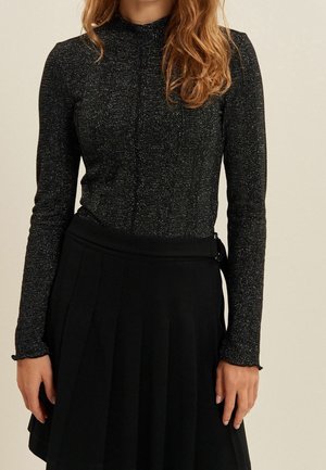 Femme portant un haut noir scintillant à manches longues rentré dans une jupe plissée noire avec une taille haute et un détail de boucle sur le côté.