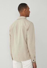 Beige, leichte Jacke mit einem kragenloser Ausschnitt, langen Ärmeln, Knopfdetails an den Manschetten und einem entspannten Schnitt. Glatte Textur, minimales Design.