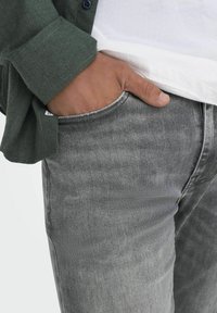 Jeans in denim grigio con leggera sbiaditura, tessuto testurizzato e dettaglio delle tasche sfilacciato. Abbinati a una camicia verde, che mette in risalto uno stile a strati.