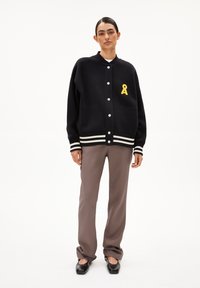 Zwarte cardigan met witte strepen, knoopsluiting aan de voorkant en voorkanten zakken. Bevat een gele lintlogo aan de linkerkant. gedragen met grijze broeken.