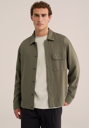 Homme porte une veste boutonnée vert olive sur une chemise blanche texturée et un pantalon noir, debout devant un fond clair uni.