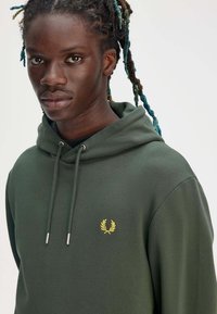 Felpa verde realizzata in morbido tessuto, con cappuccio regolabile, tasca centrale e un piccolo logo dorato ricamato sul petto.