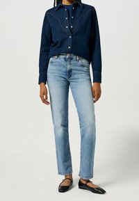 Camisa de mezclilla azul oscuro con cierre de botones y costuras en contraste, combinada con jeans de pierna recta azul claro y bailarinas negras.