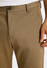 Pantaloni marroni dalla texture, con chiusura a bottone, dettagli di cucitura visibili e tasche frontali, realizzati in un morbido tessuto misto.