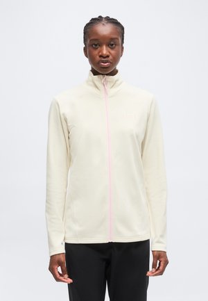 Femme aux cheveux tressés portant une veste crème à fermeture éclair intégrale avec une fermeture éclair rose clair et un pantalon noir, debout devant un fond uni.