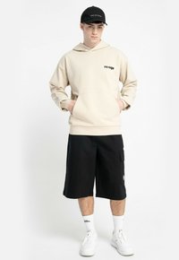 Młody mężczyzna nosi beżowy hoodie "ravage", czarne szorty cargo, białe sneakersy, białe skarpetki oraz czapkę w kolorze czarnym z napisem "MADE FOR STORMS", stoi z rękami w kieszeniach.