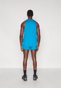 Ένας άνδρας στέκεται πλάτη, φορώντας ένα στενό μπλε αμάνικο πάνω μέρος και αντίστοιχα σορτς. Το ύφασμα φαίνεται λείο και αθλητικό, συνδυασμένο με σκούρες κάλτσες και παπούτσια.