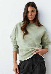 Gaiši zaļš kokvilnas džemperis ar apaļu kakla izgriezumu un ribotiem aproču galiem, kombinēts ar tumšām svītrainām biksēm. Gluda tekstūra, vaļīga piegriezuma.