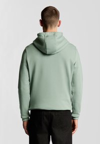 Felpa verde chiaro con cappuccio regolabile, texture liscia e polsini a costine, abbinata a pantaloni scuri, vista da dietro.