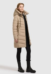 Beige gewatteerde jas met een hoge kraag en grote capuchon, voorzien van een ritssluiting aan de voorkant, zijzakken en een doorgestikte textuur. Draagt bij zwarte leggings en laarzen.