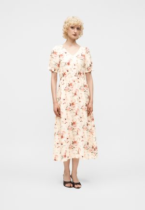 Kvinde står iført en blomsterprint off-white midikjole med puffede korte ærmer og sorte åbne hæle mod en ensfarvet lys baggrund.