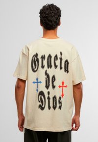 Camiseta de algodón en color beige con mangas cortas. Presenta letras negras "Gracia de Dios" y diseños de cruces rojas y azules en la parte posterior.