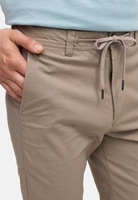 Beige puuvillahousut, joissa on naru vyötäröllä, sileä tekstuuri ja sivutaskut, joissa on ommeltu vyötärö ja kontrastiväriset narun päät.