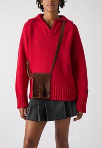 Röd stickad tröja med V-ringning och ribbad nederdel, i kombination med en brun lädercrossbody-väska och en plisserad svart rutiga kjol.