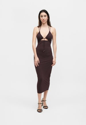 Γυναίκα με μακριά σκούρα μαλλιά που φοράει ένα μίντι μωβ φόρεμα χωρίς μανίκια με κοψίματα και μαύρα ψηλοτάκουνα σανδάλια, στέκεται μπροστά σε λευκό φόντο.