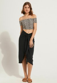 Haut court évasé à épaules nues avec un motif géométrique noir et blanc, associé à une jupe noire nouée. Des sandales en toile marron complètent le look.