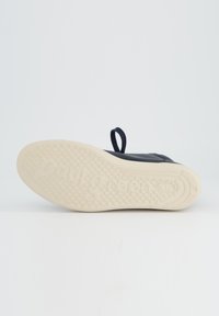 Semelle de chaussure noire avec un caoutchouc blanc texturé pour une meilleure traction, arborant le nom de la marque en relief. Fini lisse avec une languette à l'arrière.