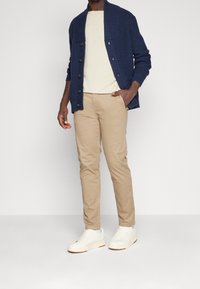 Man i marinblå stickad cardigan, beige byxor, krämfärgad t-shirt och vita sneakers står med en hand i fickan.