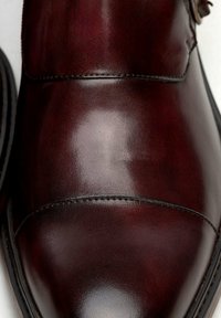 Zapato de cuero marrón con un acabado brillante, que presenta acentos cosidos y una puntera redondeada. El diseño incluye una textura suave y un hardware mínimo.