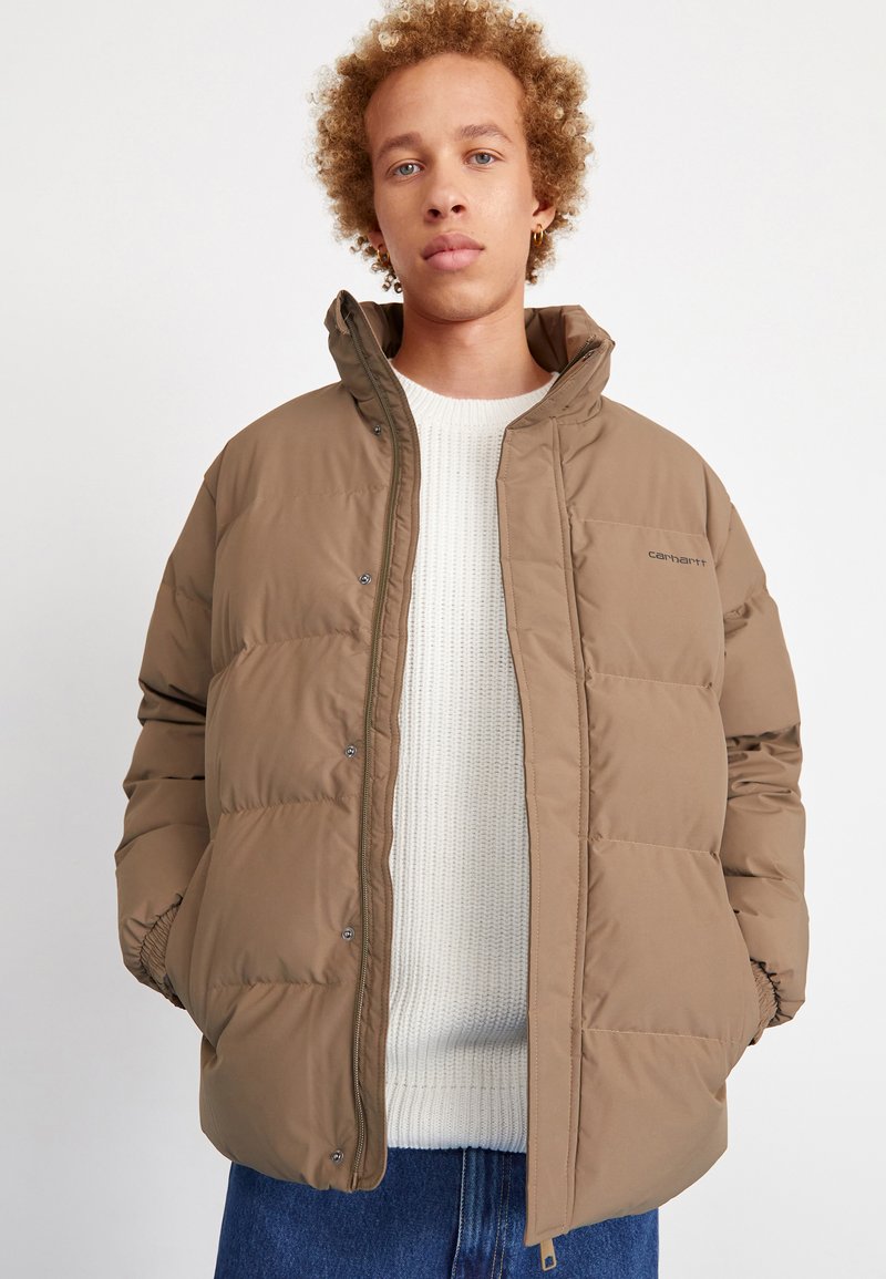 Brown puffer jacket with snap buttons, elastic cuffs, and side pockets. Worn over a light beige knit sweater, featuring a relaxed fit.