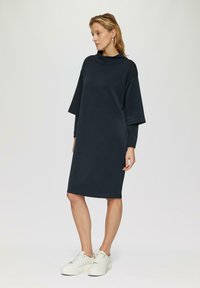 Donkerblauwe jurk met een hoge halslijn, oversized mouwen en een ontspannen pasvorm. Gecombineerd met witte sneakers voor een casual look.