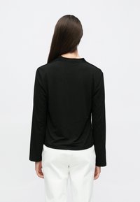 Melnas krāsas garām piedurknēm apģērbts tops ar apaļu kakla izgriezumu, gludu tekstūru un nelielu kabatas drapējumu aizmugurē, savienots ar baltām biksēm.