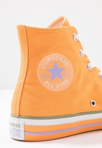 Kraťasová oranžová plátěná teniska s bílými očky, kulatou nášivkou s fialovou hvězdou a duhovou proužkovanou podrážkou.